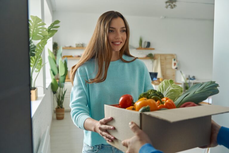 Happy young woman accepting box with groceries from delivery man at home