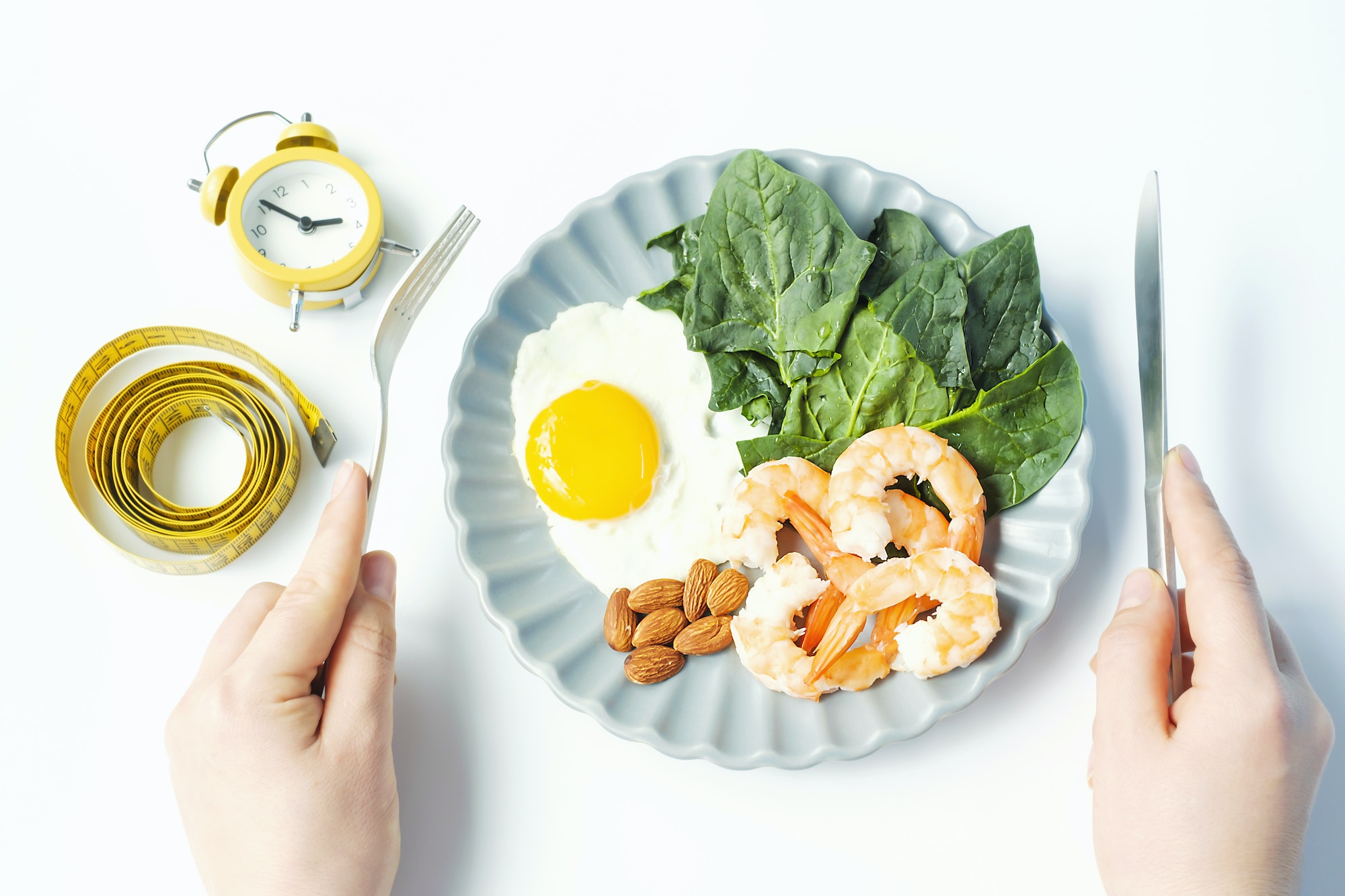Female hands holding a fork with a knife next to plate of food on a white background top view.